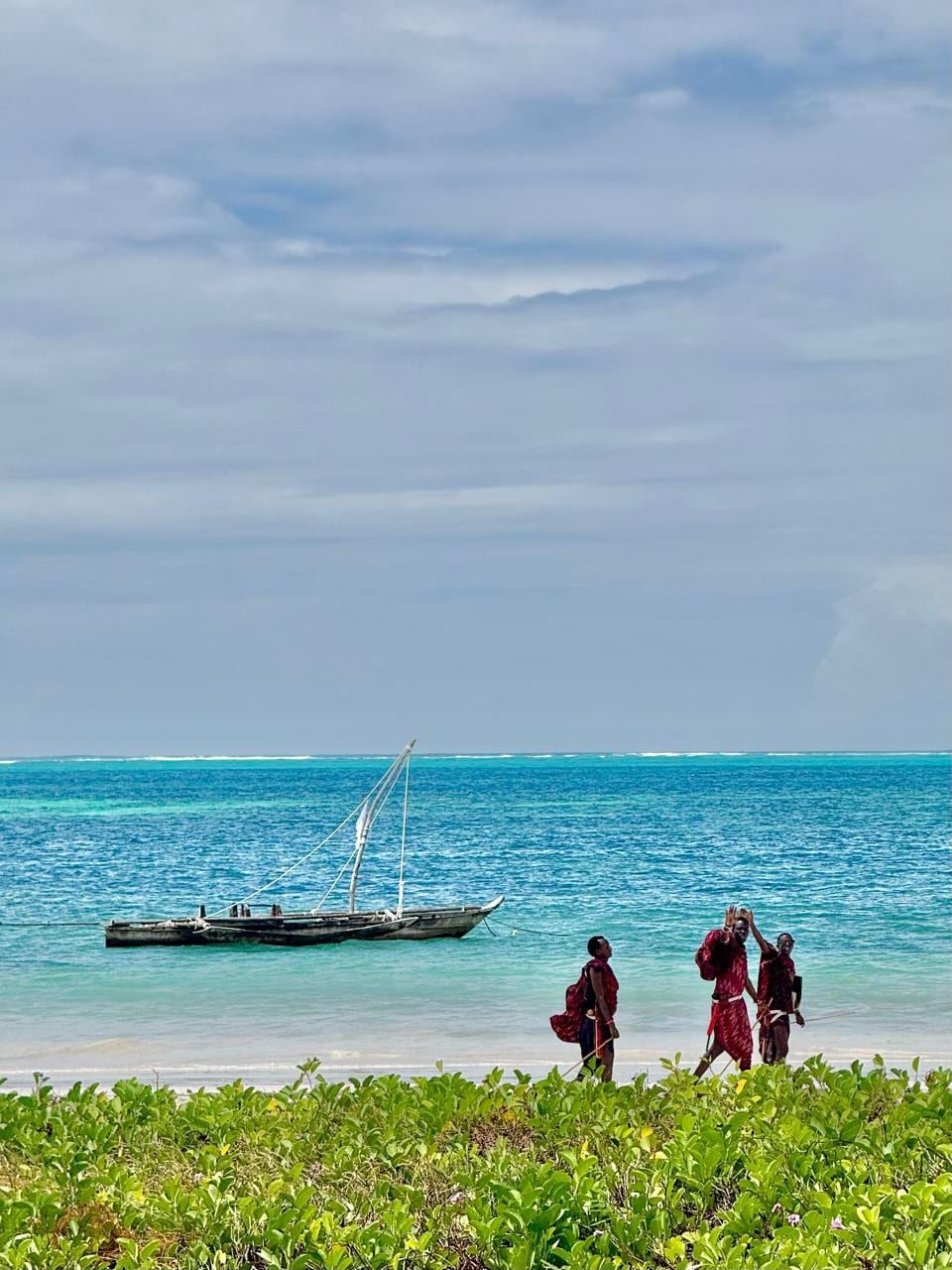 Three Maasai in red walking the shoreline past a moored dhow, Zanzibar