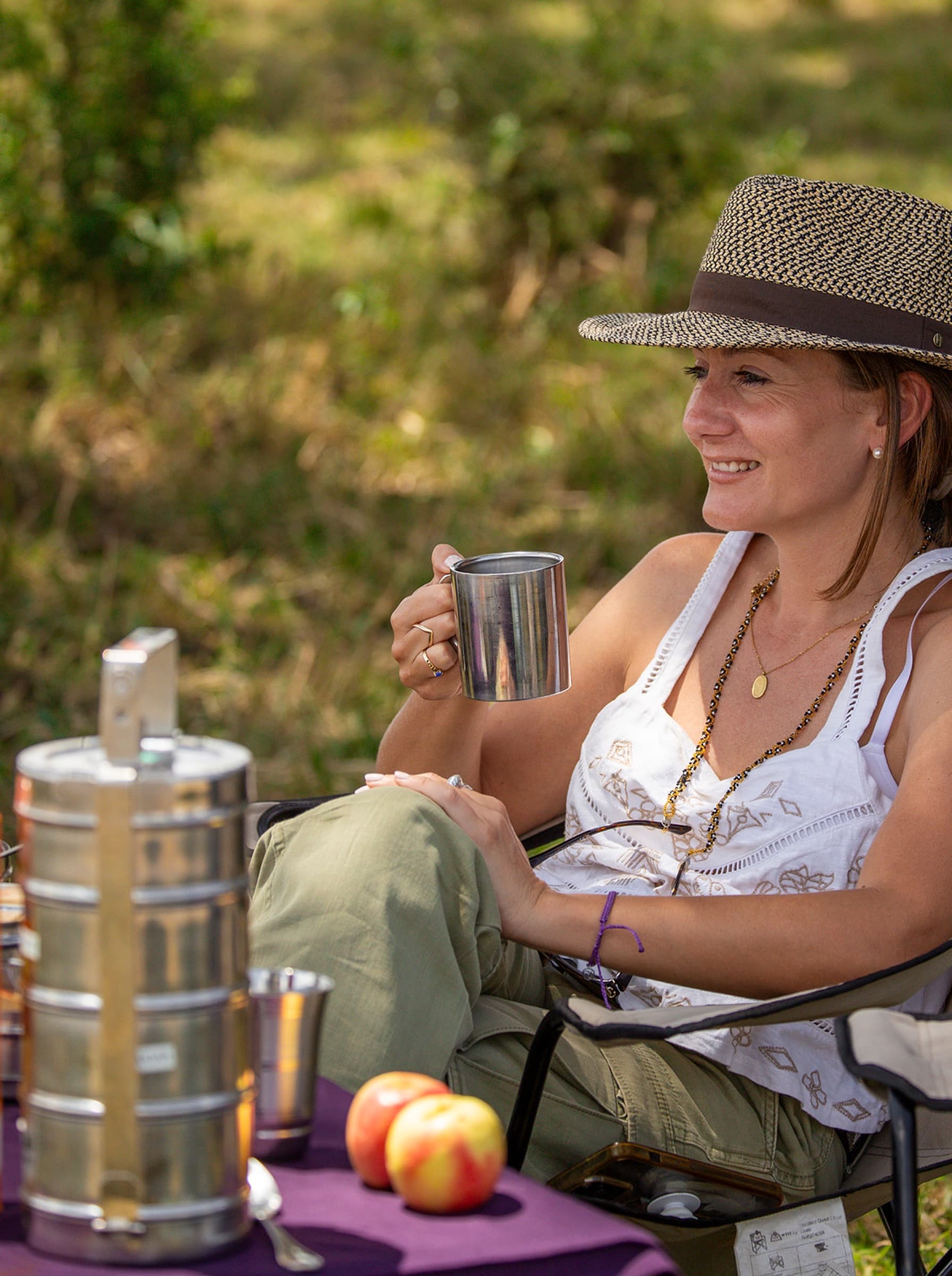 Danielle in a camp chair with a tin mug of coffee, hat on, laughing