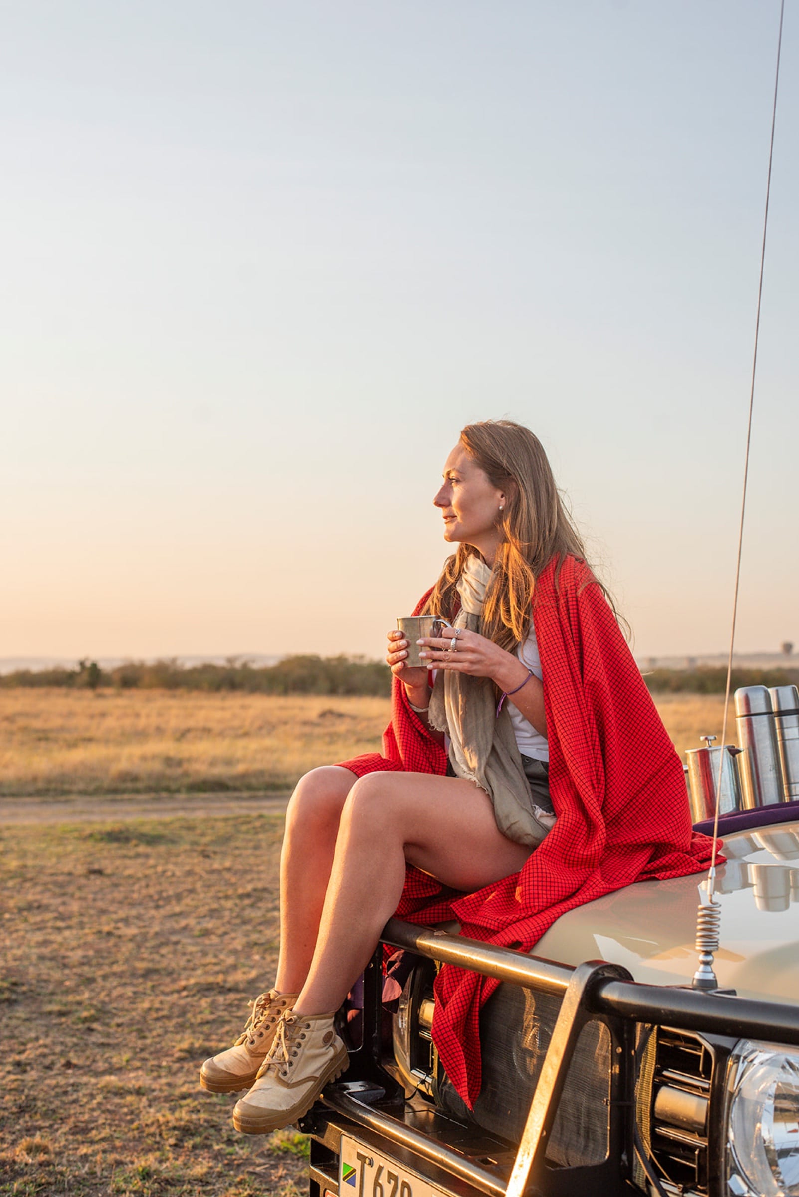 Danielle wrapped in a red blanket on the bonnet of a safari vehicle at sunrise, mug in hand