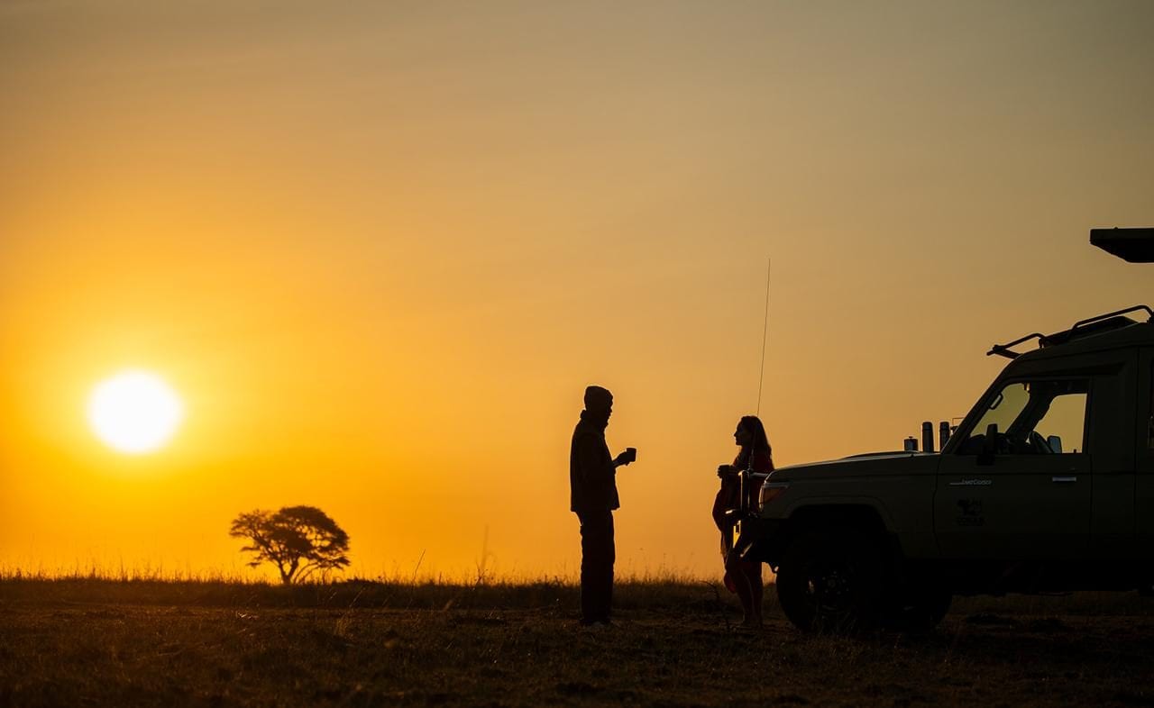 Danielle and a guide with coffee beside a safari vehicle, silhouetted against the rising sun