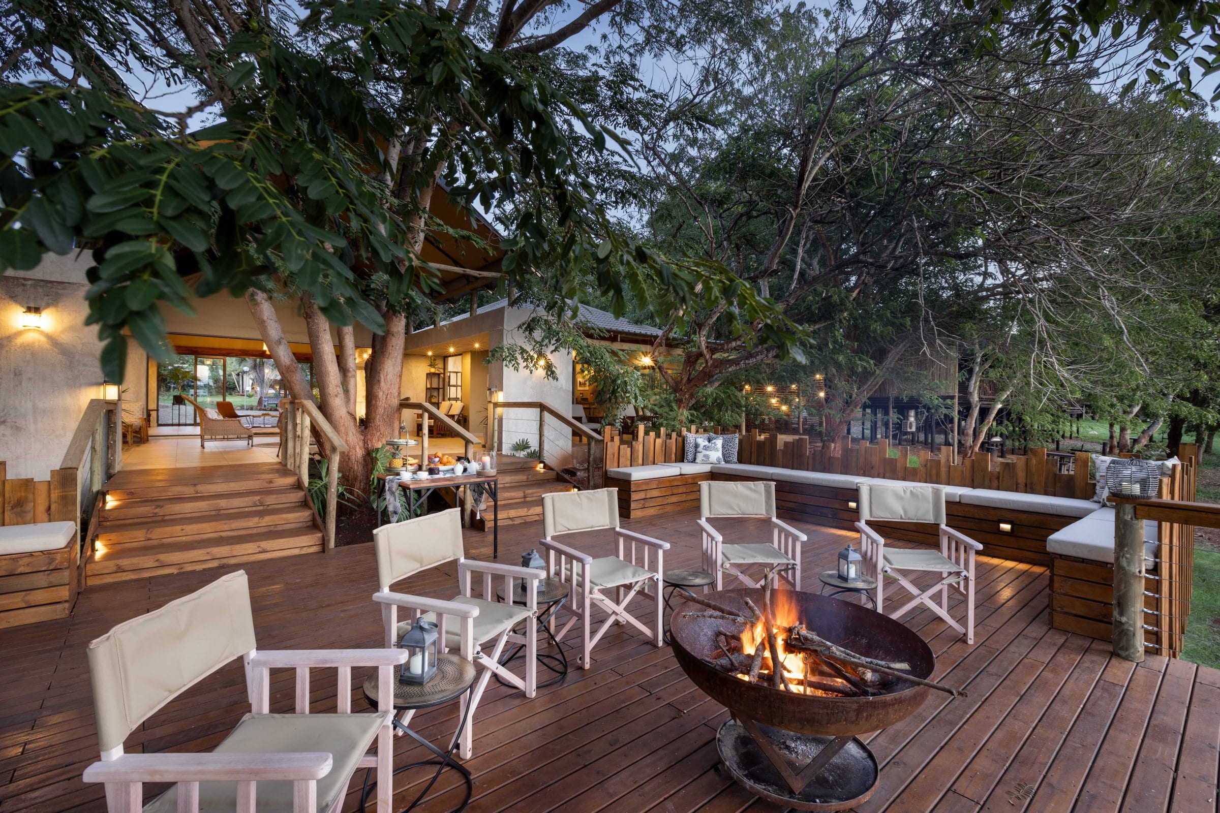 The camp deck at dusk, canvas chairs around a fire bowl under big trees