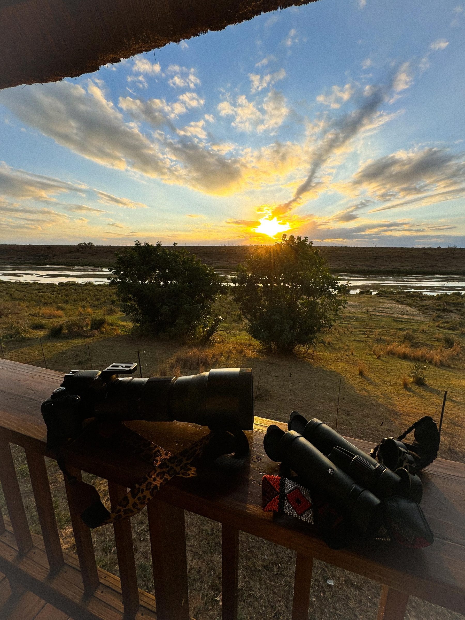 A camera with a long lens and binoculars on the deck rail, the sun setting over the river