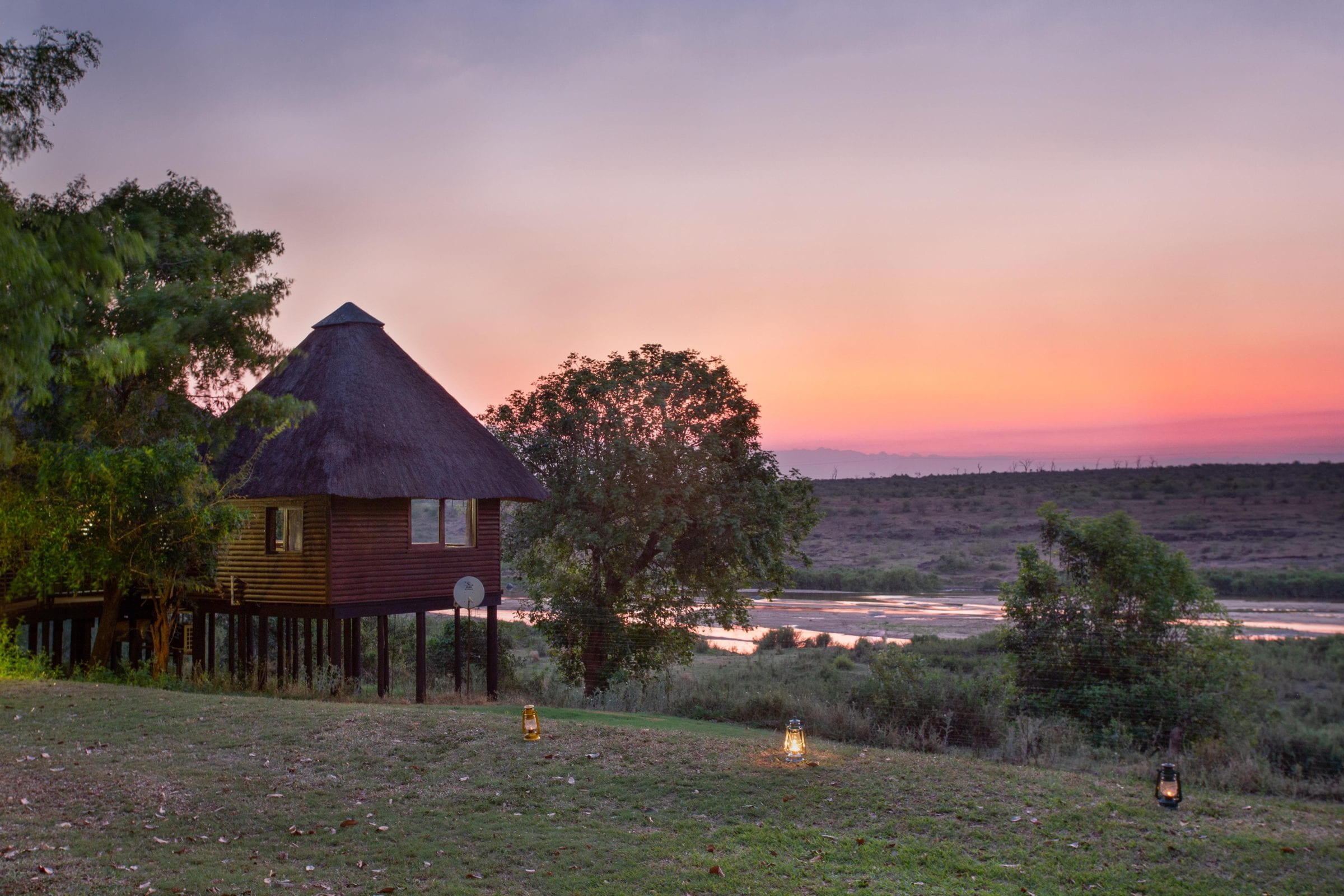 A thatched treehouse on stilts at sunset, lanterns lit on the lawn