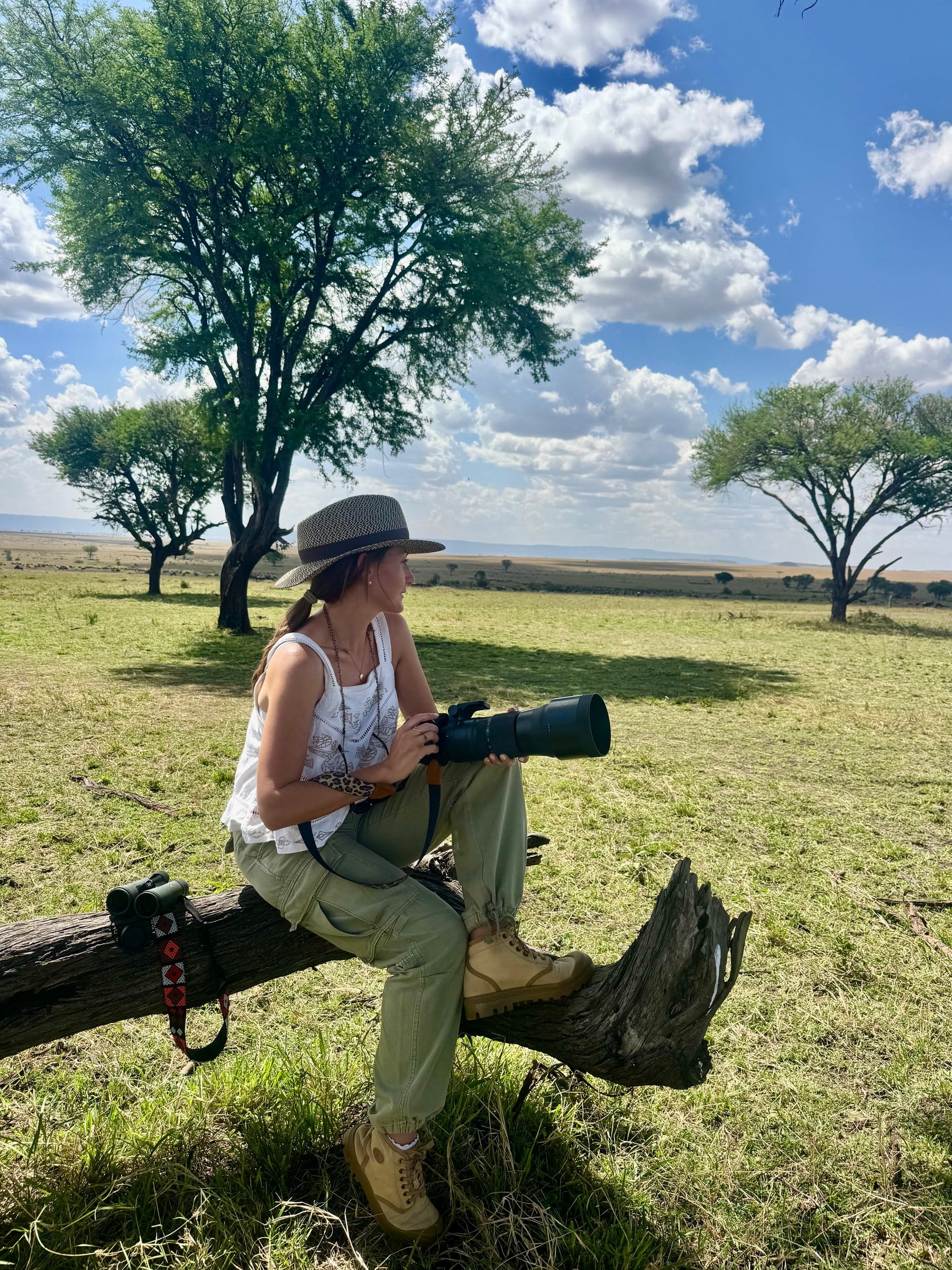 Danielle sitting on a fallen log with a long camera lens, an open plain and acacias behind her